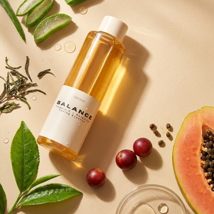 A clear bottle of Fabyoulife BALANCE Organic Balance Cleansing & Exfoliating Serum & Elixir with a white label, styled in a flat-lay with green tea leaves, sliced aloe vera, papaya, and small red berries on a beige background.