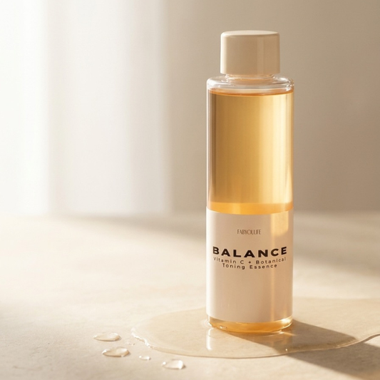 A bottle of FABYOULIFE Balance Vitamin C & Botanical Toning Essence with a white cap, filled with a golden-amber liquid, standing on a light beige surface with a small puddle of liquid around its base.